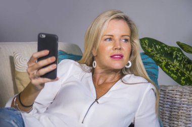 güzel ve mutlu sarışın kadın erken 40s evde oturma odası koltuk kanepe yaşam tarzı kavram rahat yatan gülümseyen cep telefonunda internet sosyal medya app kullanarak rahat