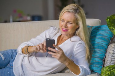 güzel ve mutlu sarışın kadın erken 40s evde oturma odası koltuk kanepe yaşam tarzı kavram rahat yatan gülümseyen cep telefonunda internet sosyal medya app kullanarak rahat