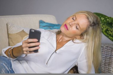 güzel ve mutlu sarışın kadın erken 40s evde oturma odası koltuk kanepe yaşam tarzı kavram rahat yatan gülümseyen cep telefonunda internet sosyal medya app kullanarak rahat