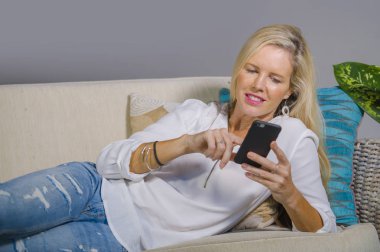 güzel ve mutlu sarışın kadın erken 40s evde oturma odası koltuk kanepe yaşam tarzı kavram rahat yatan gülümseyen cep telefonunda internet sosyal medya app kullanarak rahat