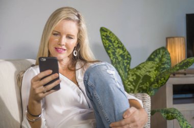 güzel ve mutlu sarışın kadın erken 40s evde oturma odası oturma rahat kanepe kanepede yaşam tarzı kavram gülümseyen cep telefonunda internet sosyal medya app kullanarak rahat