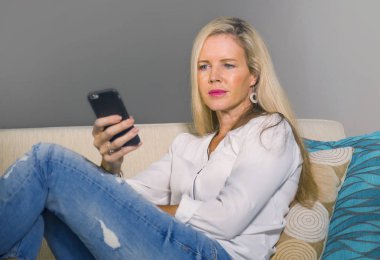 güzel ve mutlu sarışın kadın erken 40s evde oturma odası oturma rahat kanepe kanepede yaşam tarzı kavram gülümseyen cep telefonunda internet sosyal medya app kullanarak rahat
