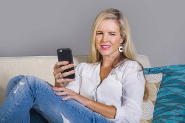 güzel ve mutlu sarışın kadın erken 40s evde oturma odası oturma rahat kanepe kanepede yaşam tarzı kavram gülümseyen cep telefonunda internet sosyal medya app kullanarak rahat
