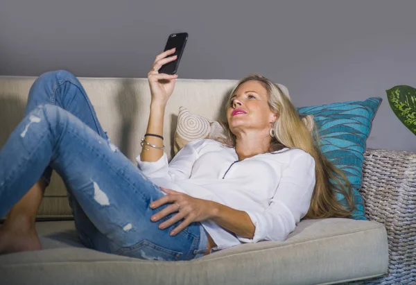 güzel ve mutlu sarışın kadın erken 40s evde oturma odası koltuk kanepe yaşam tarzı kavram rahat yatan gülümseyen cep telefonunda internet sosyal medya app kullanarak rahat