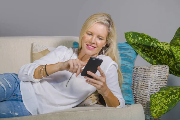 güzel ve mutlu sarışın kadın erken 40s evde oturma odası koltuk kanepe yaşam tarzı kavram rahat yatan gülümseyen cep telefonunda internet sosyal medya app kullanarak rahat