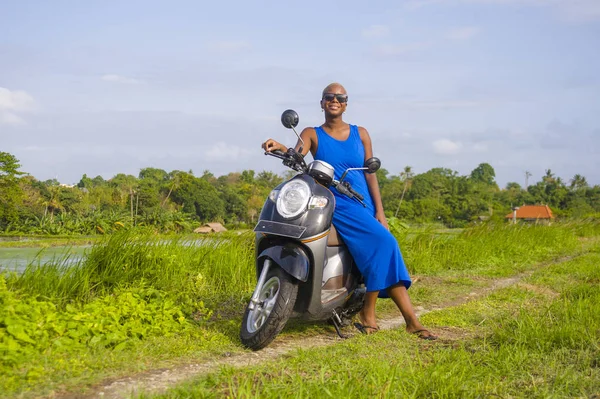 young beautiful and happy african american black woman posing cool on scooter motorbike at green field tropical landscape in adventure trip and summer holidays travel concept