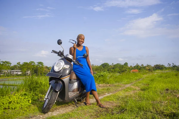 young beautiful and happy african american black woman posing cool on scooter motorbike at green field tropical landscape in adventure trip and summer holidays travel concept