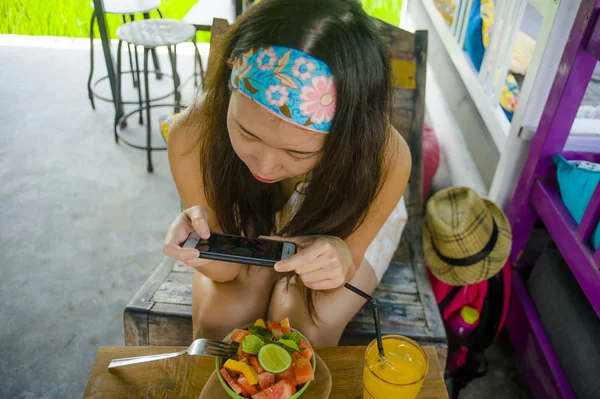 young happy and pretty digital nomad Asian Korean woman taking picture of fruit salad with mobile phone for sharing on internet social media in healthy nutrition and travel blogger food review