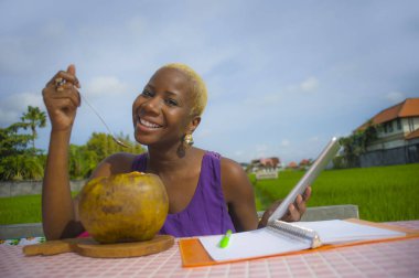 Açık havada içme hindistan cevizinin içine dijital göçebe başarı ve online serbest iş tropikal yerde dijital tablet pad ile çalışan genç çekici ve mutlu başarılı siyah afro Amerikan kadın