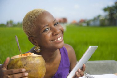 Açık havada içme hindistan cevizinin içine dijital göçebe başarı ve online serbest iş tropikal yerde dijital tablet pad ile çalışan genç çekici ve mutlu başarılı siyah afro Amerikan kadın