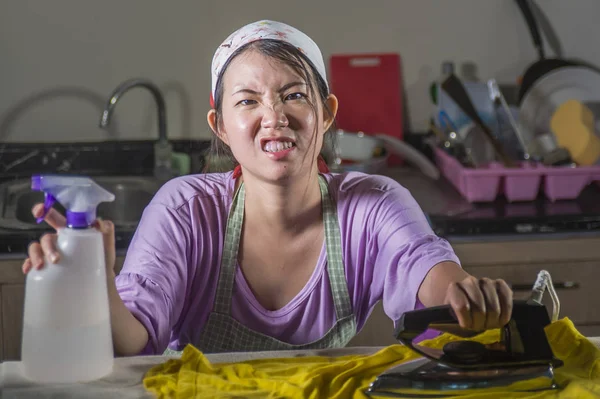 young pretty frustrated and stressed Asian Chinese woman with iron working at home kitchen ironing clothes desperate and overwhelmed in house maid service housework domestic stress