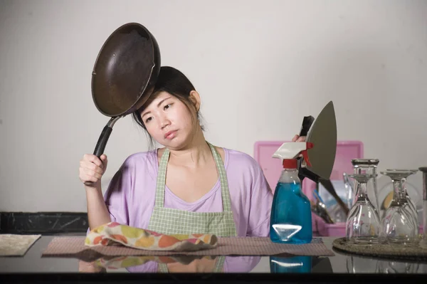 young beautiful and frustrated Asian Korean woman holding pan and iron stressed and upset overwhelmed by domestic work and chores at home kitchen in housekeeping stress concept