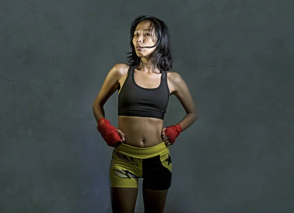 portrait of sporty and fit fighter Asian Chinese woman using wrist wraps training mma fight sport or boxing workout looking dangerous in badass pose isolated on dark grunge background