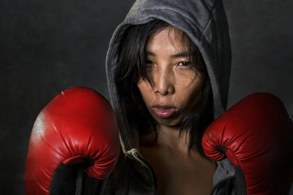 close up portrait of young fit and healthy Asian Korean boxer woman in fitness top hoodie and boxing gloves posing cool badass attitude angry and defiant in fight workout isolated on dark