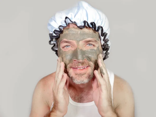 lifestyle funny portrait of happy weird man on shower cap looking to himself in bathroom mirror with green cream on his face applying facial mask skin care product smiling cheerful and fresh