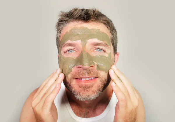 lifestyle isolated portrait of young attractive and happy man looking to himself in bathroom mirror with green cream on his face applying facial mask skin care product smiling cheerful and fresh