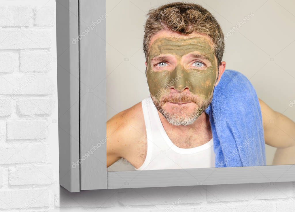 lifestyle funny portrait of young attractive and surprised man horrified looking himself on bathroom mirror ugly and weird applying green cream on his face in facial mask male beauty product concept