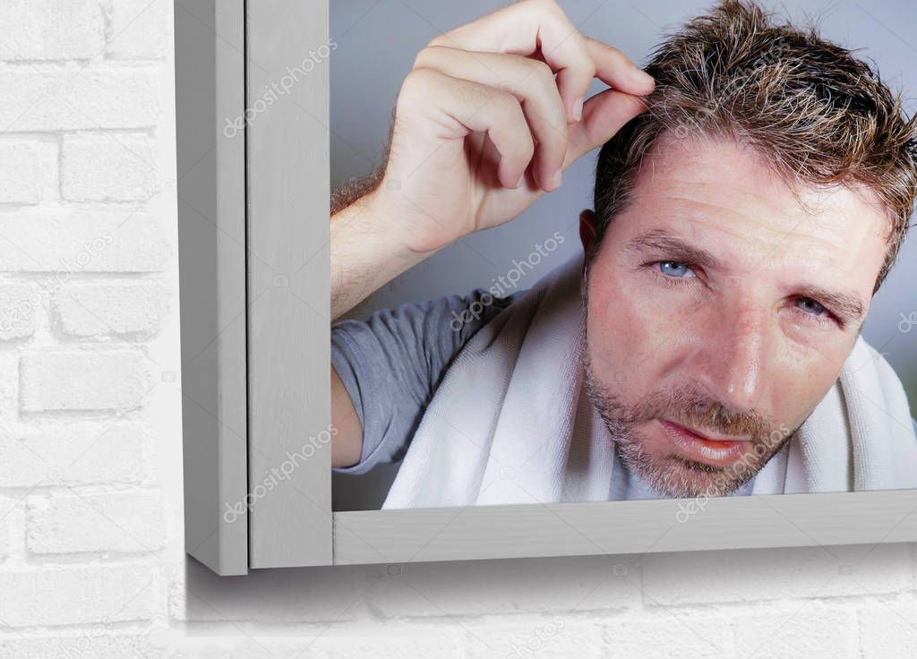 lifestyle portrait of attractive worried and concerned Caucasian man looking at bathroom mirror finding himself with gray hair in head feeling unhappy aging getting grey hairs and getting older