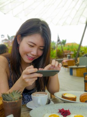 Genç güzel ve mutlu Asya Kore seyahat blogger kadının fotoğrafını cep telefonu, açık havada kahve dükkanı gülümseyen güzel kahvaltı tablo ile sosyal medya gıda yazı kavram olarak memnun