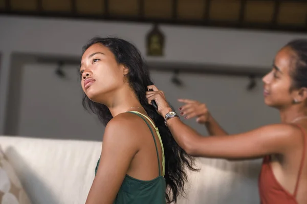 young beautiful and happy Asian girlfriends at home couch with one girl brushing the hair of the other woman helping preparing for dating smiling cheerful and relaxed together