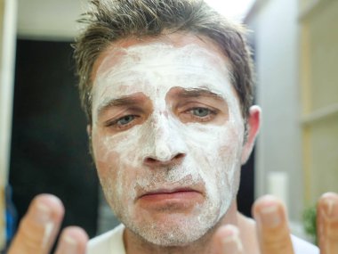 Genç komik çekici adam ev banyo tuvalet aynası sağlamlılık yüzündeki kendini tuhaf ve çirkin beyaz maske ile erkek güzellik cilt bakım ve hijyen kavramında bulma uygulama bakarak