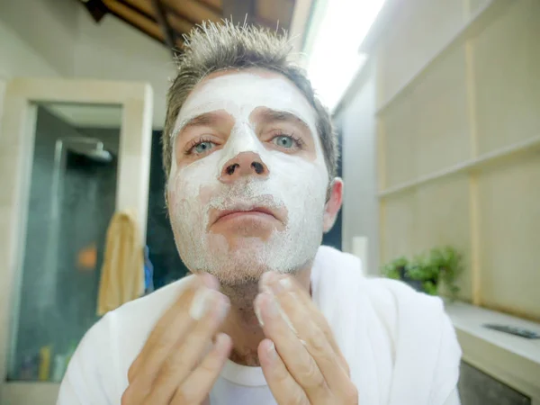 close up face portrait of young handsome and attractive Caucasian man at home looking at himself on bathroom mirror applying white facemask in male beauty and facial mask hygiene concept