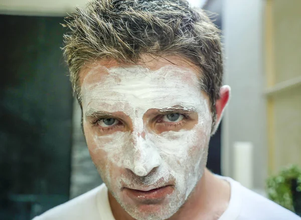 close up face portrait of young handsome and attractive Caucasian man at home looking at himself on bathroom mirror with facemask in male beauty and facial mask hygiene concept