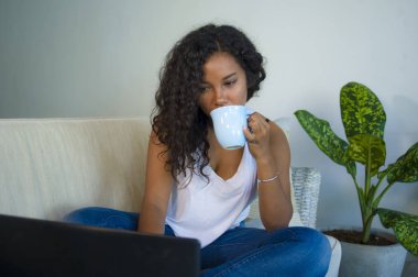 yaşam tarzı portre genç çekici ve rahat siyah afro Amerikan öğrenci kadın kanepe evde oturan içme kahve veya çay kot pantolon ve singlet giyen dizüstü bilgisayarınızla ağ kanepe