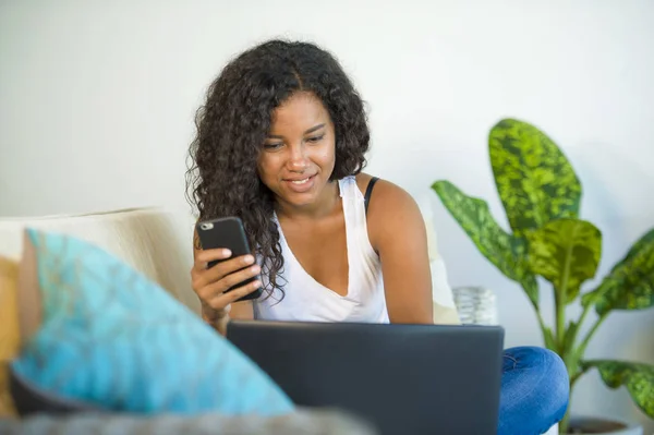 lifestyle portrait of young happy and beautiful black african American woman using internet  mobile phone while working on laptop computer at home living room sofa couch relaxed