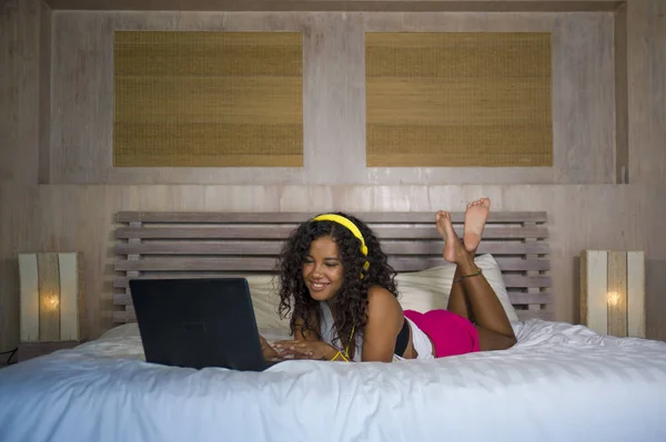 lifestyle portrait of young beautiful and happy black African American woman at home bedroom lying cheerful on bed listening to internet music with yellow headphones and laptop computer smiling