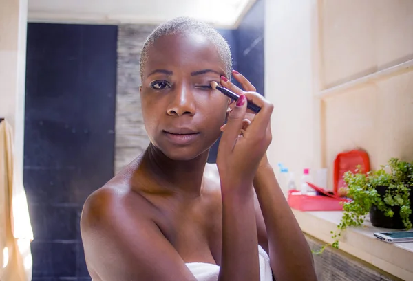 lifestyle natural portrait of young attractive and happy black afro American woman at home bathroom applying face makeup with brush looking at toilet mirror fresh in beauty cosmetics concept