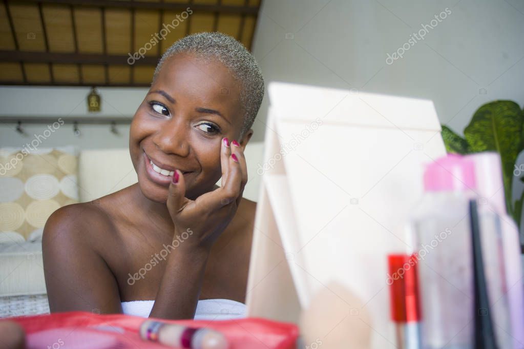 young happy and attractive black african American woman wrapped in towel applying makeup cosmetics using fingers putting base primer looking and smiling to the mirror in female beauty concept