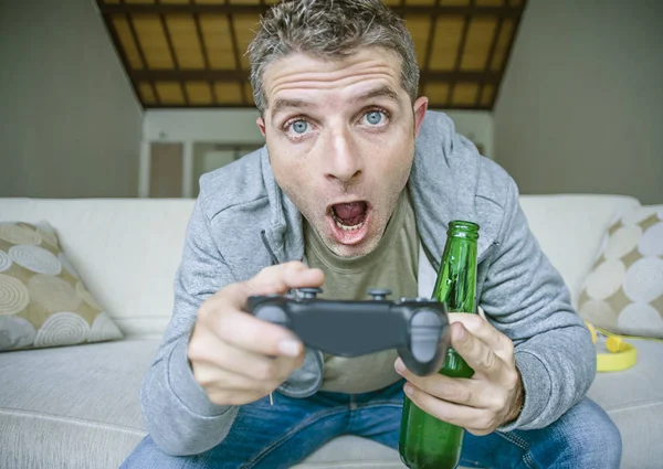 lifestyle portrait of young happy and excited gamer man playing video game at home drinking beer having fun on sofa couch holding controller stressed and concentrated in online  gaming concept