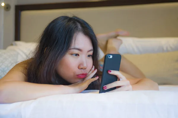 lifestyle portrait of young happy and pretty Asian Korean woman at home bedroom or hotel room lying on bed relaxed smiling cheerful using internet online dating app texting on mobile phone