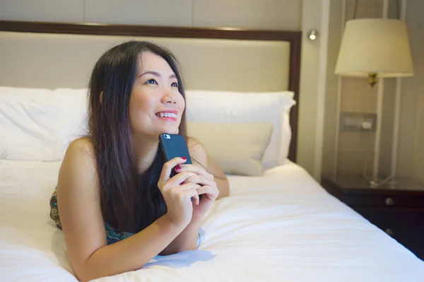 lifestyle portrait of young happy and pretty Asian Korean woman at home bedroom or hotel room lying on bed relaxed smiling cheerful using internet online dating app texting on mobile phone