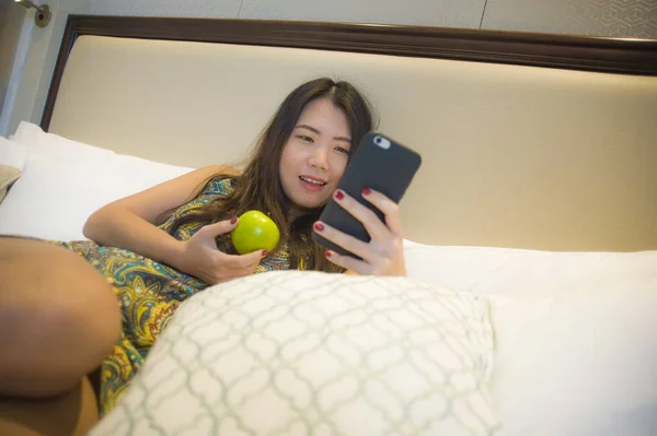 年轻快乐和漂亮的亚洲韩国妇女的生活方式肖像放松在家庭卧室或酒店房间躺在床上吃苹果使用互联网在线约会应用程序发短信手机 — 图库照片