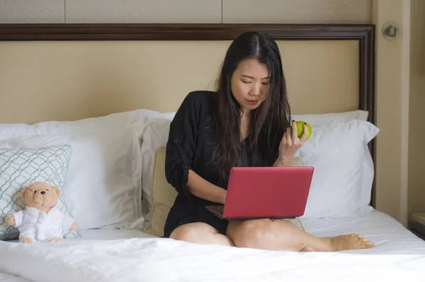 lifestyle portrait of young happy and relaxed Asian Korean woman at hotel room bed working with laptop computer eating apple cheerful in business travel and successful entrepreneur concept