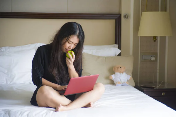 年轻快乐和放松的生活方式肖像在酒店房间床上的亚洲华人妇女与笔记本电脑一起吃苹果愉快的商务旅行和成功的企业家理念 — 图库照片