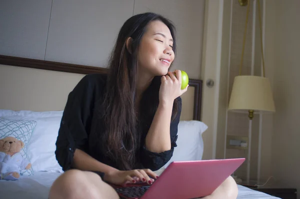 青年快乐和放松的生活方式肖像在酒店房间床上的亚洲韩国妇女与笔记本电脑一起吃苹果愉快的商务旅行和成功的企业家理念 — 图库照片