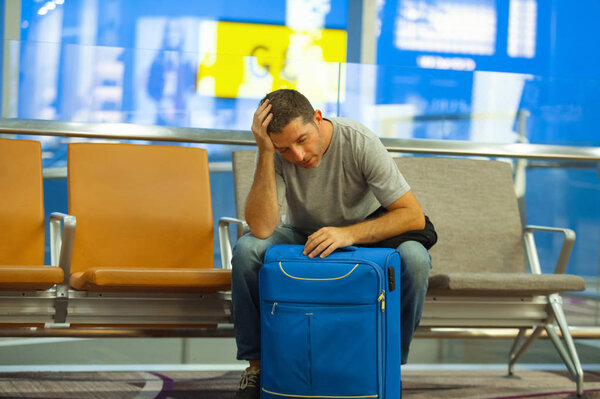 lifestyle portrait in airport of young attractive and tired tourist man with suitcase sitting worried and stressed at boarding gate waiting for cancelled or delayed flight ready for holidays travel