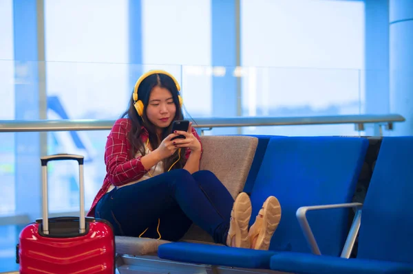 lifestyle portrait of young happy and pretty Asian Korean tourist woman sitting at airport departure boarding gate waiting for flight using internet on mobile phone smiling in holidays travel