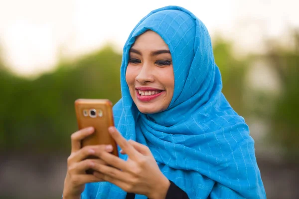 lifestyle isolated portrait of young happy and beautiful tourist woman in muslim hijab head scarf using mobile phone outdoors smiling cheerful in holidays travel and smartphone technology concept