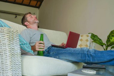 yaşam tarzı kapalı genç çekici ve yakışıklı mutlu adamın evde online dizüstü bilgisayar netbook kullanarak çalışma koltuk kanepe oturma portre rahat rahat içki bira şişesi 