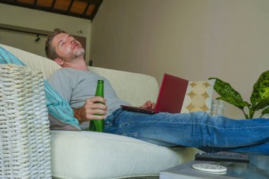 yaşam tarzı kapalı genç çekici ve yakışıklı mutlu adamın evde online dizüstü bilgisayar netbook kullanarak çalışma koltuk kanepe oturma portre rahat rahat içki bira şişesi 