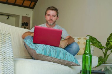 yaşam tarzı kapalı dizüstü bilgisayar netbook kullanarak çevrimiçi çalışıyorsanız koltuk kanepe evde oturan genç çekici ve yakışıklı mutlu bir adam portresi bira şişesi ile rahat rahat 