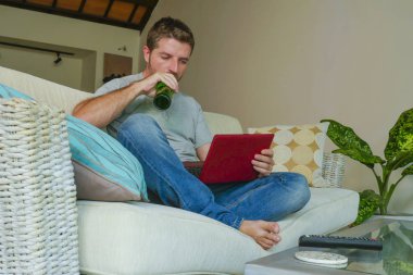 yaşam tarzı kapalı genç çekici ve yakışıklı mutlu adamın evde online dizüstü bilgisayar netbook kullanarak çalışma koltuk kanepe oturma portre rahat rahat içki bira şişesi 