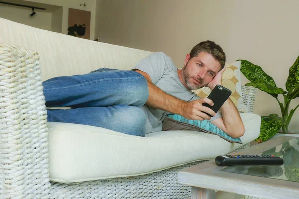 lifestyle indoors portrait of young handsome and attractive happy man using internet dating online app on mobile phone or networking from home sofa couch with smartphone relaxed