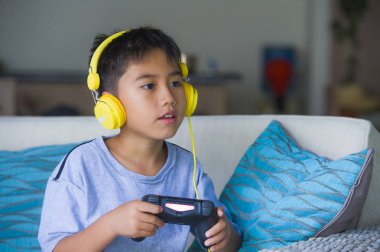 Genç İspanyol küçük çocuk heyecanlı ve mutlu online video oyun denetleyicisi zevk sahip holding kulaklık ile oynarken portresi eğlenceli oyun duygu ve çocuk oyun bağımlılığı kanepede oturan
