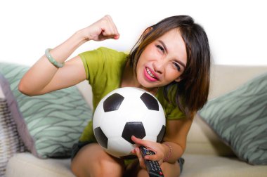 lifestyle portrait at home of young happy and beautiful football fan woman watching television game sitting on sofa couch holding soccer ball excited celebrating scoring goal and victory clipart