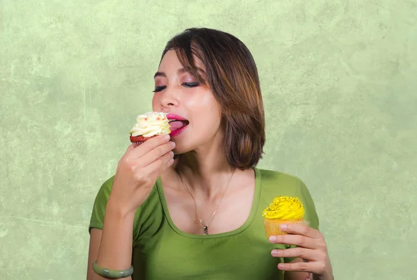 young beautiful and happy hispanic woman eating tasty and delicious yellow sugary cupcakes posing on isolated background in sugar and sweet addiction and unhealthy nutrition concept